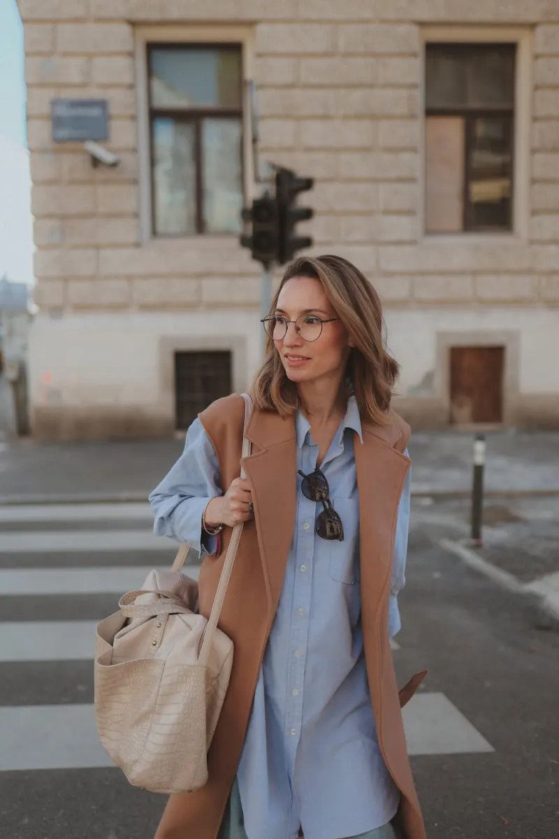 Žena u plavoj košulji i prsluku stoji na ulici i smiješi se